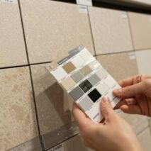 A person holding a grout colour swatch card against a beige tile to find a matching grout shade