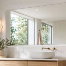 Large frameless bathroom mirror above a modern vanity with warm timber cabinetry, white tiles and natural light